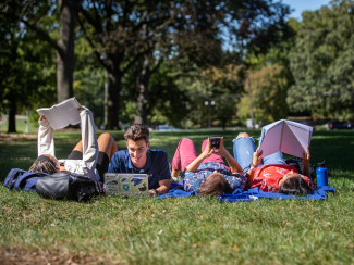Four students laying in the grass each viewing a book, laptop, or phone.