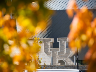 A view of the UK logo made out of stone at the end of a wall framed by out of focus leaves in fall.