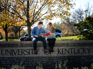 A male and female student reviewing a red notebook while sitting on a stonewall engraved with University of Kentucky.