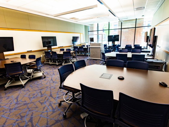 An empty classroom with multiple display monitors mounted at groupings of tables and chairs.
